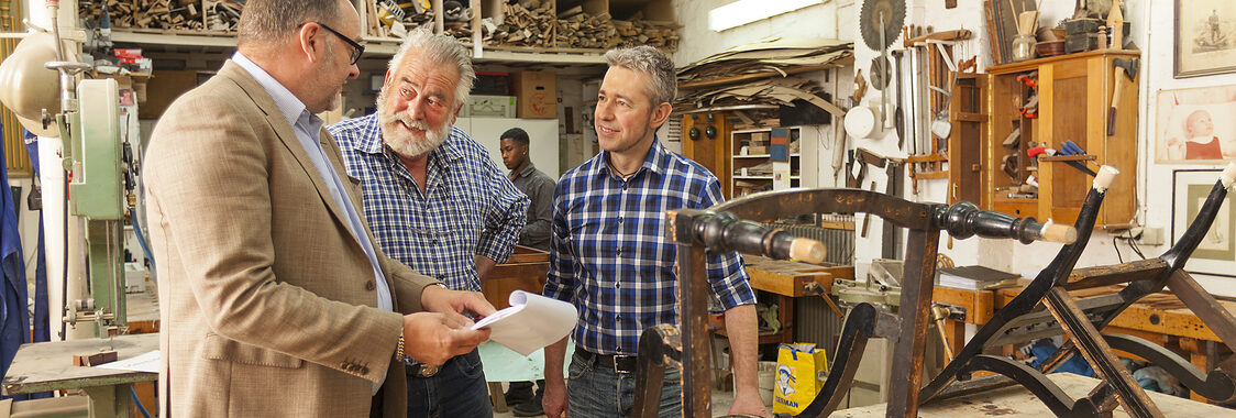 Ein Berater mit Jacket spricht in einer Tischler-Werkstatt mit zwei Handwerkern