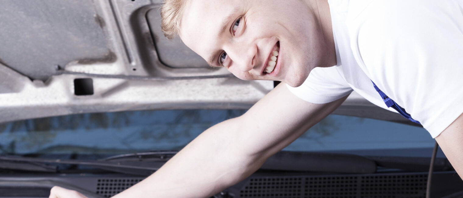 Ein junge Mann ist über den Motorraum eines Fahrzeugs gebeut. Er hat den Kopf in Richtung Kamera gedreht und lächelt.