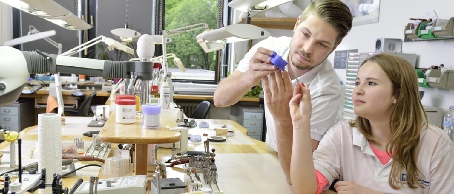 In einer Werkstatt sehen sich ein junger Mann und eine junge Frau einen blauen Zahnabdruck an. Die Frau hält ein Werkzeug.