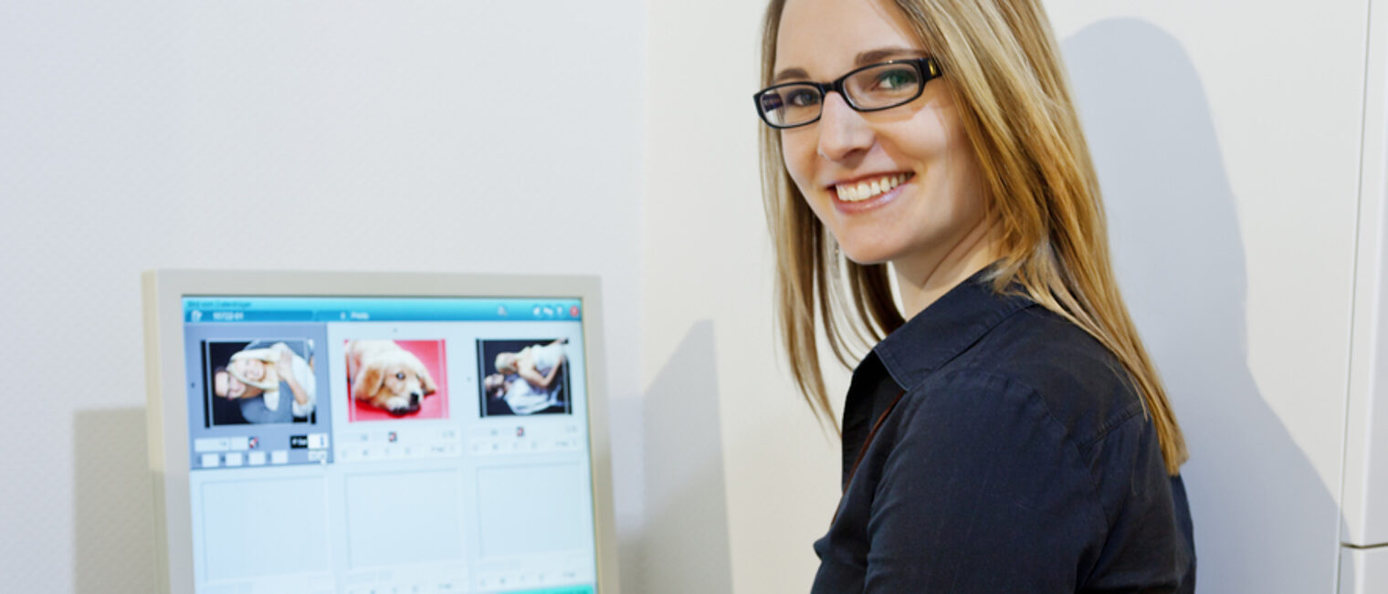 Blonde Frau mit Brille sitzt vor einem Bildschirm, auf dem drei Fotos zu sehen sind. Sie hat sich halb zur Kamera umgedreht.