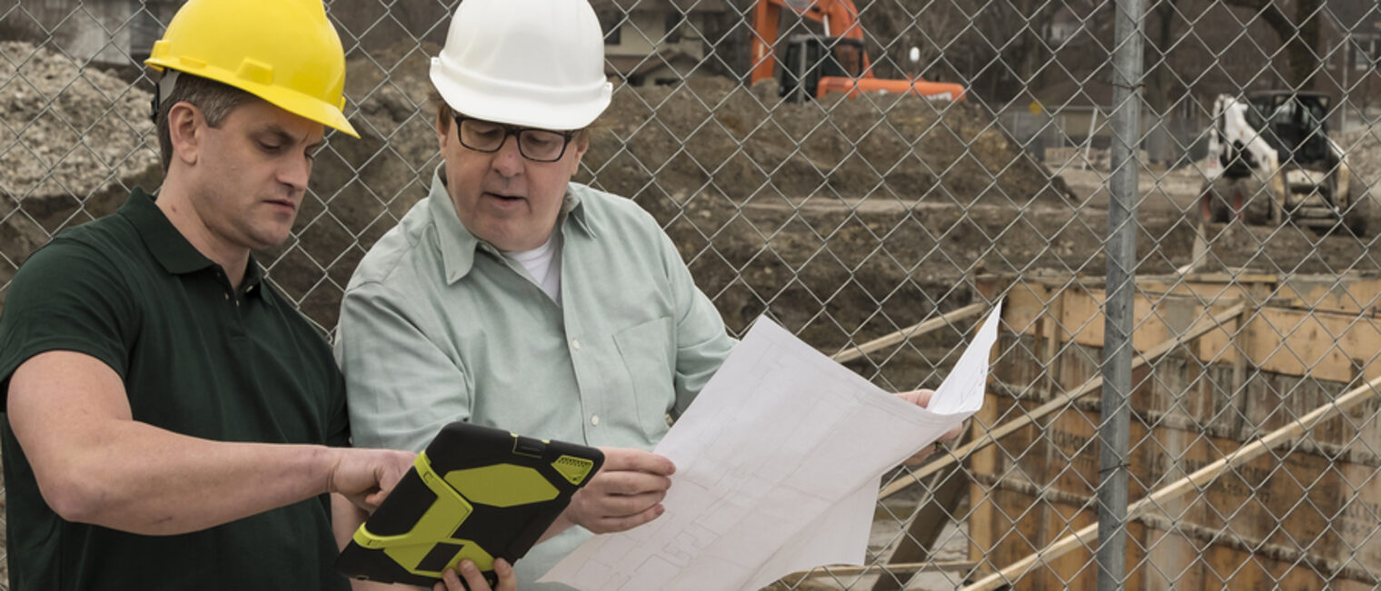 Zwei Männer mit Helmen schauen auf einen Plan. Im Hintergrund ist eine Baustelle zu sehen.