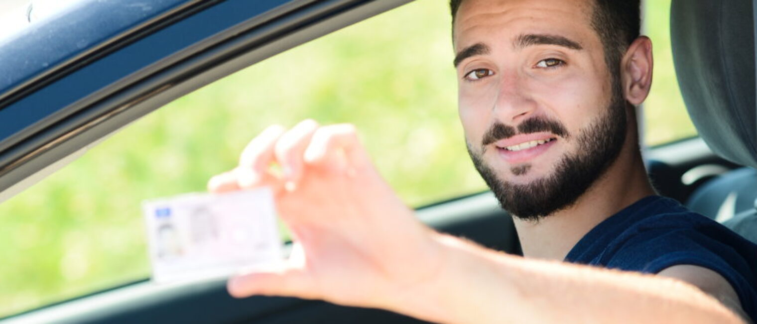 Ein junger Mann schaut aus dem geöffneten Fahrerfenster eines Pkws und hält seinen Führerschein in die Kamera.