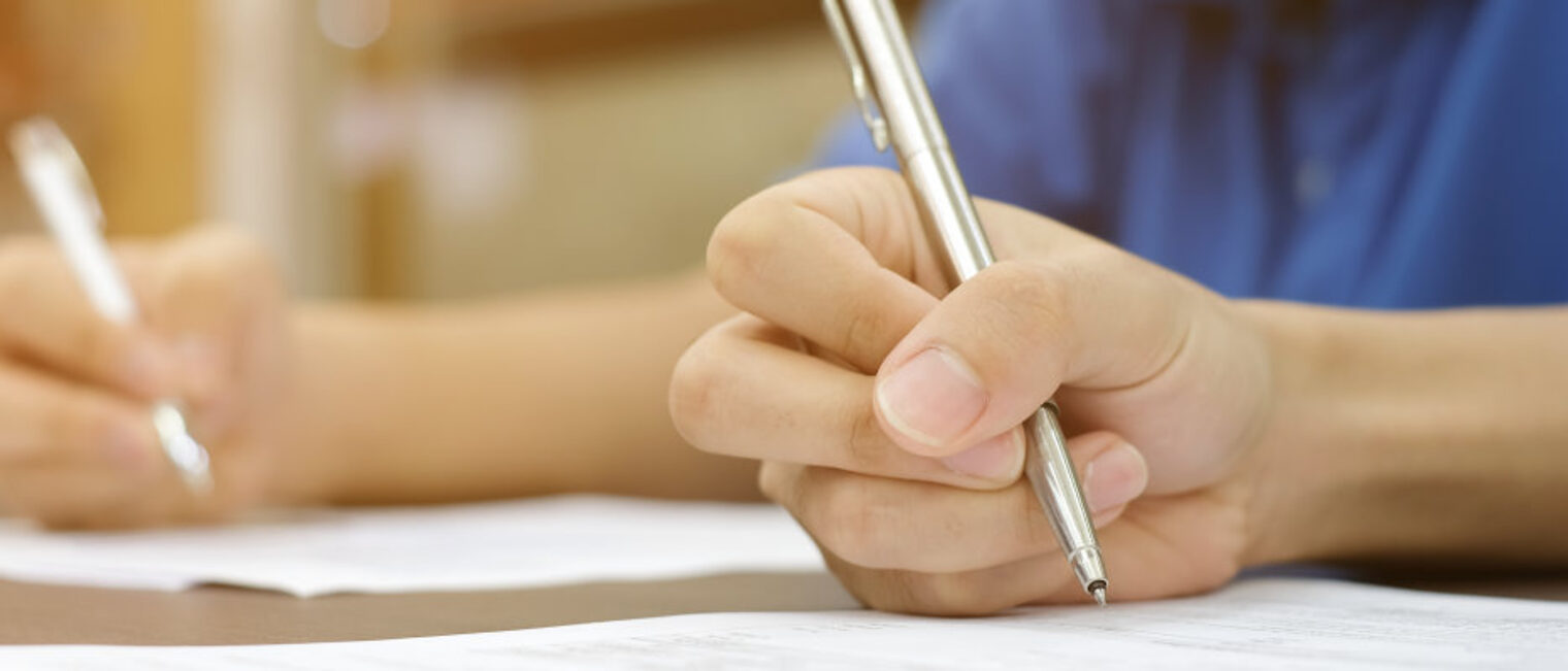 Eine Hand hält einen Stift über ein Blatt Papier auf einem Tisch. Im Hintergrund ist eine weitere Hand zu sehen.