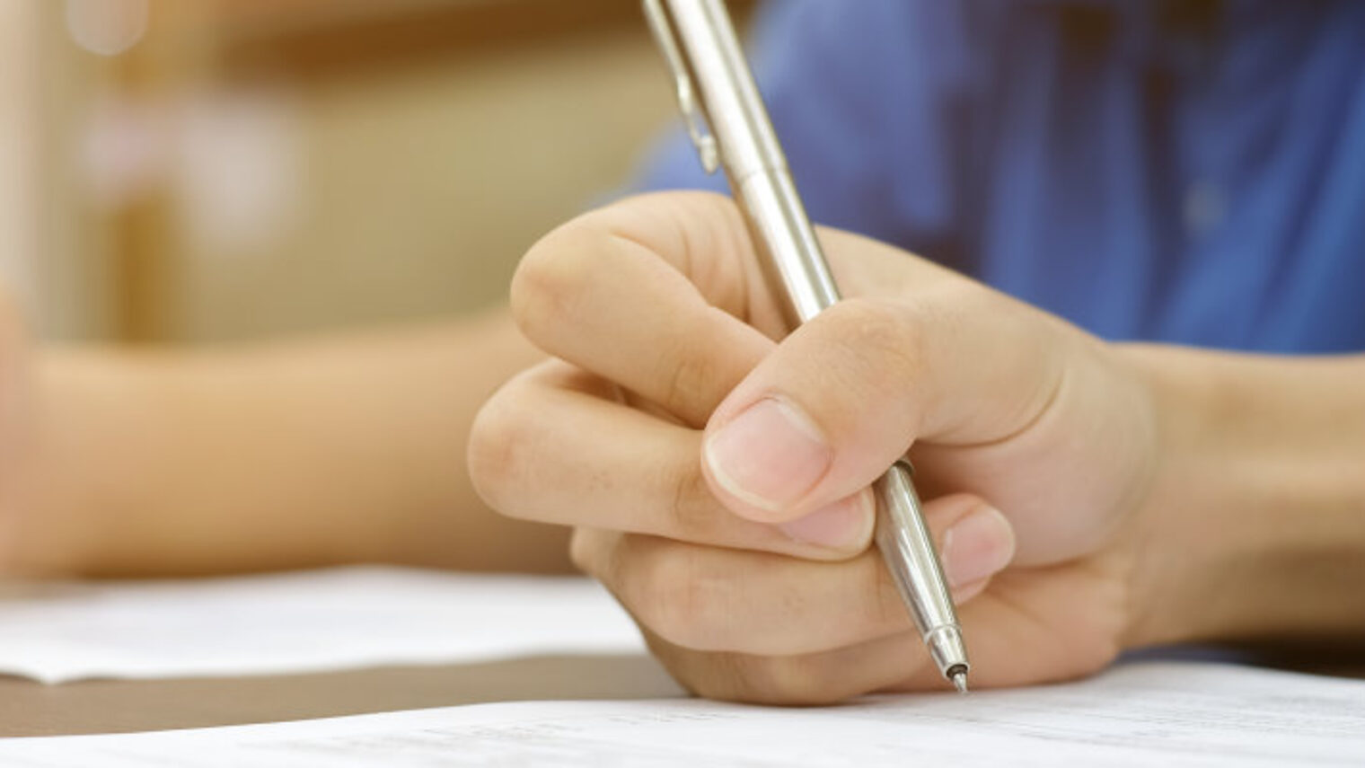 Eine Hand hält einen Stift über ein Blatt Papier auf einem Tisch. Im Hintergrund ist eine weitere Hand zu sehen.