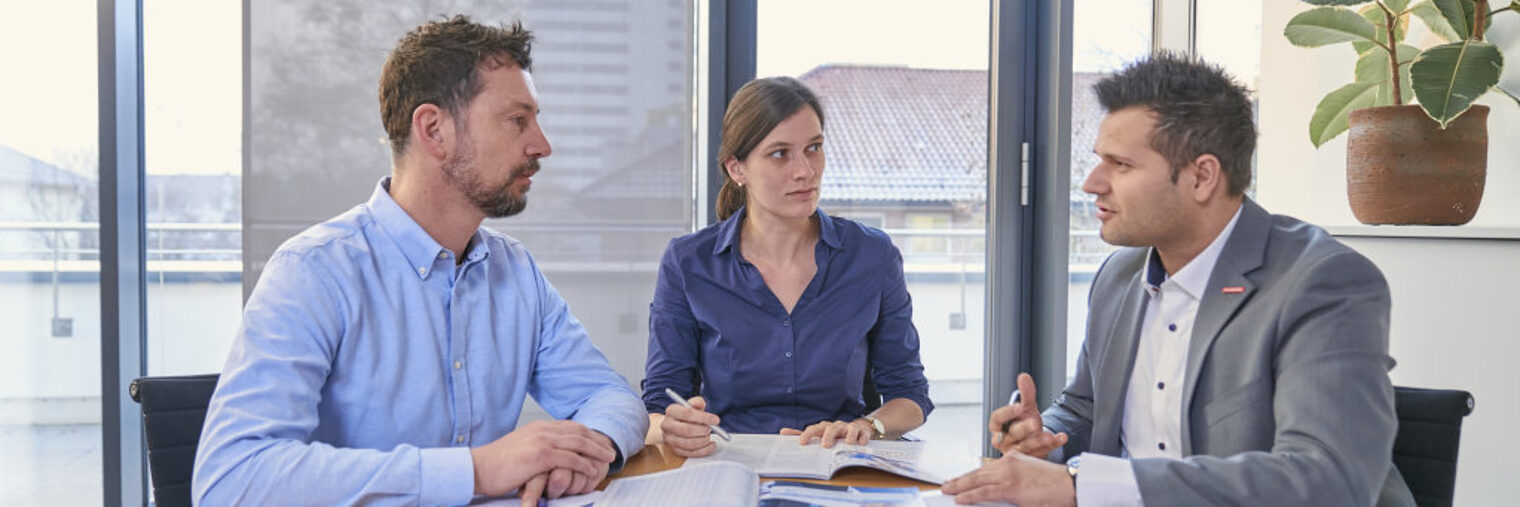 Eine Frau und zwei Männer sitzen an einem Besprechungstisch in einem freundlichen und modernen Büro
