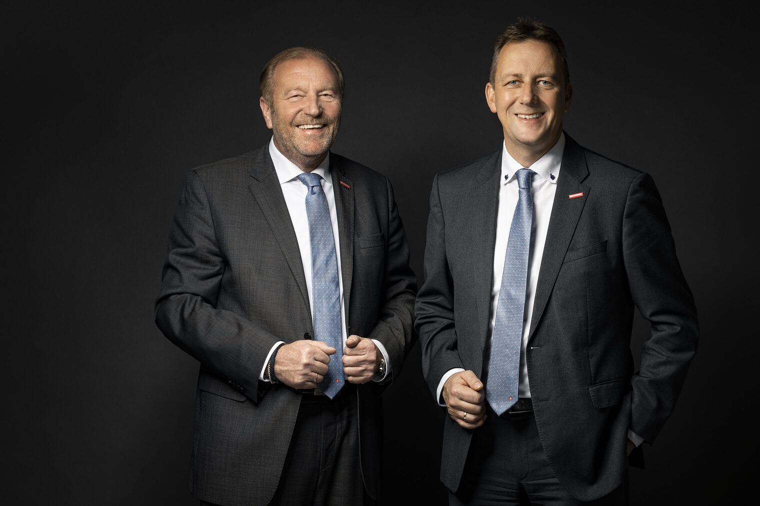 Zwei Männer in Anzügen und Krawatten stehen nebeneinander vor einem dunklen Hintergrund und lächeln in die Kamera.