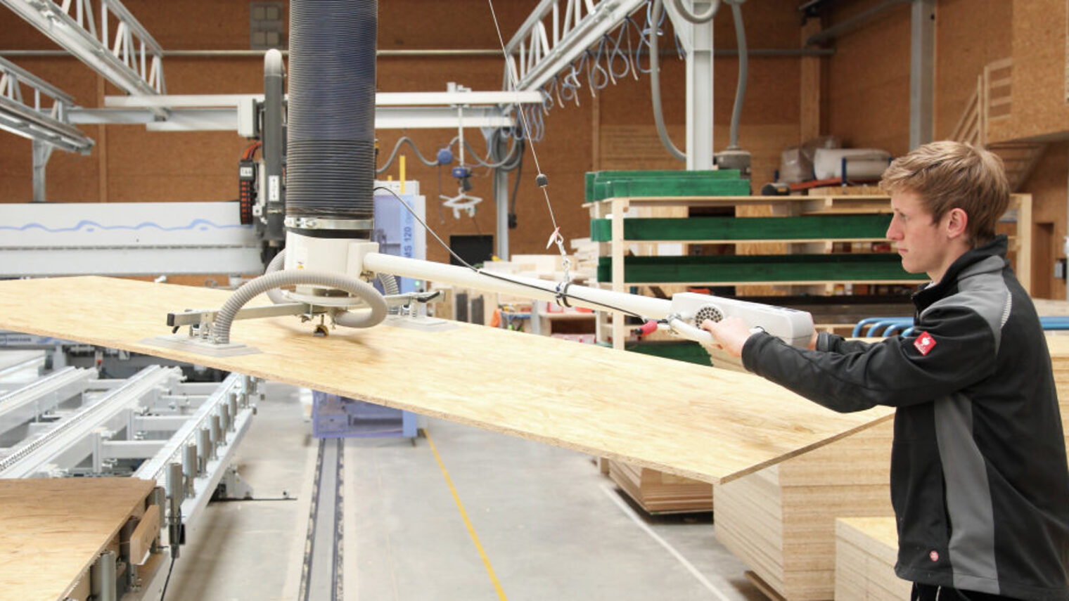 Junger Mann hebt mit Hilfe einer Maschine eine Platte aus Holz von einem Transportband in einer Fertigungshalle.