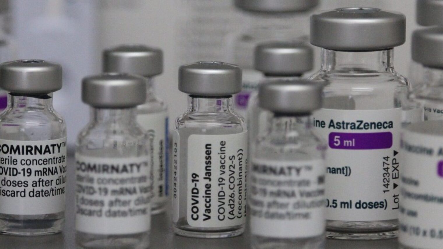 Kleine Fläschchen in verschiedenen Größen mit den Labels der Impfstoffe gegen Corona wie Comirnaty, Janssen und AstraZeneca.