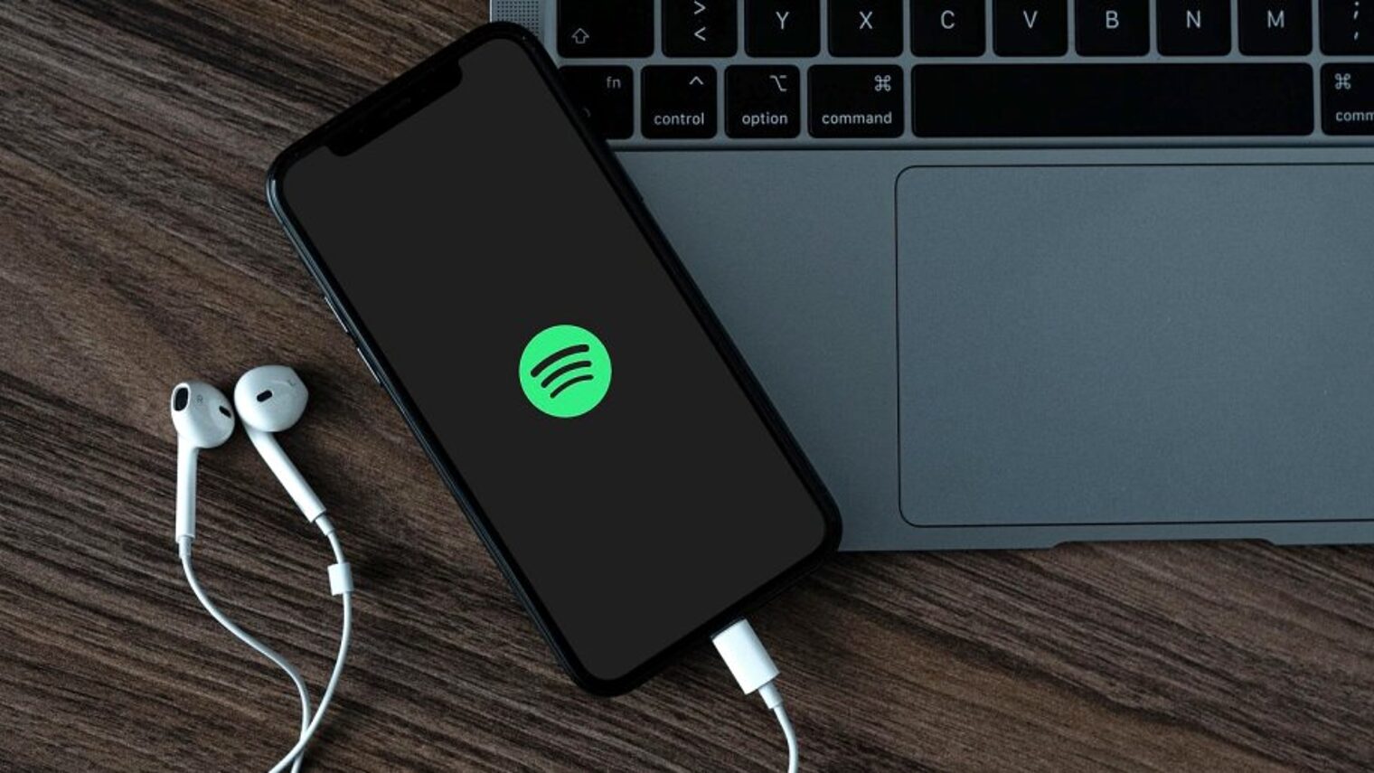 Auf einer Holzfläche steht ein Laptop, darauf ein Smartphone mit rundem grünen Symbol im Display und weißen In-Ear-Kopfhörern