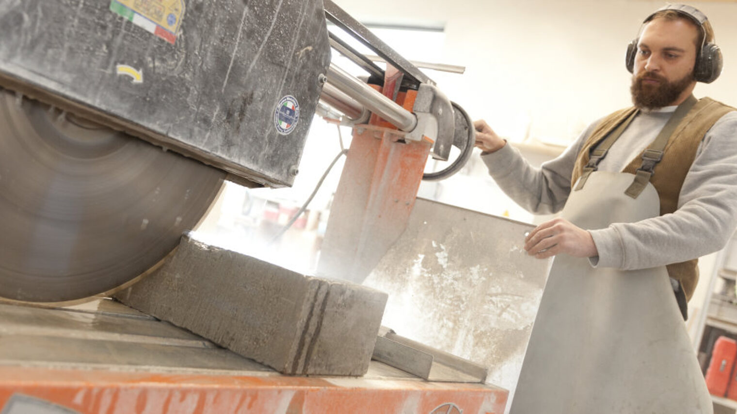 Mann in Arbeitskleidung sägt mit einer großen maschinellen Säge einen Steinblock in einer Werkstatt.