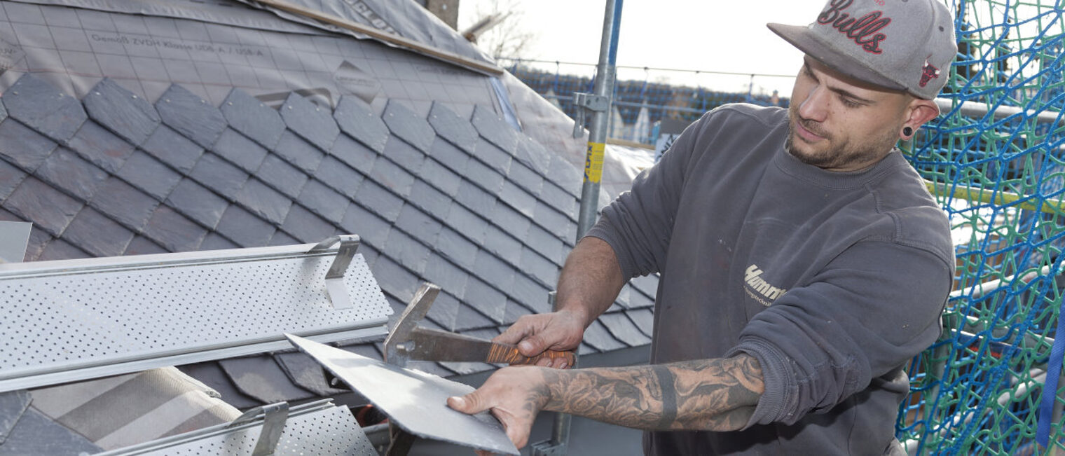 Ein Mann steht auf einem Dach und schneidet eine Dachschindel zurecht. 