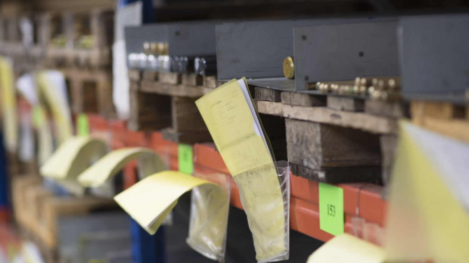 In einem Lager für Material sieht man viele Fächer auf Paletten, in denen Metallstäbe liegen. Davor gelbe Zettel und Nummern.