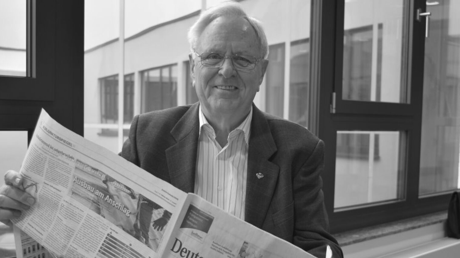 Älterer Mann mit Brille sitzt vor Fenster mit einer Zeitung in der Hand, dargestellt in schwarz-weiß.