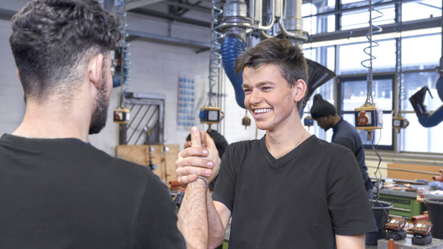 Zwei junge Männer in schwarzen T-Shirts begrüßen sich in einer Werkstatt lachend mit einem Handschlag.
