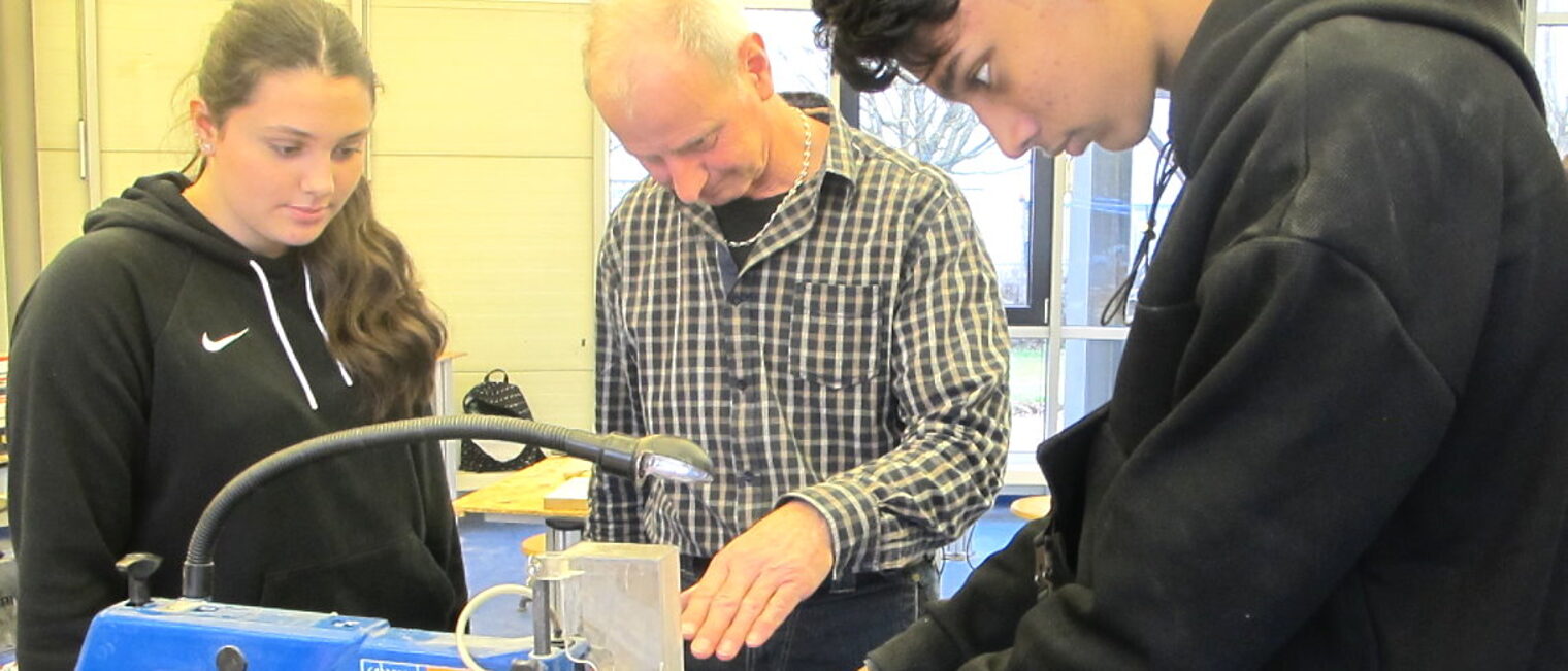 Ein Mann in der Mitte zeigt einem Schüler rechts und einer Schülerin links eine Maschine.