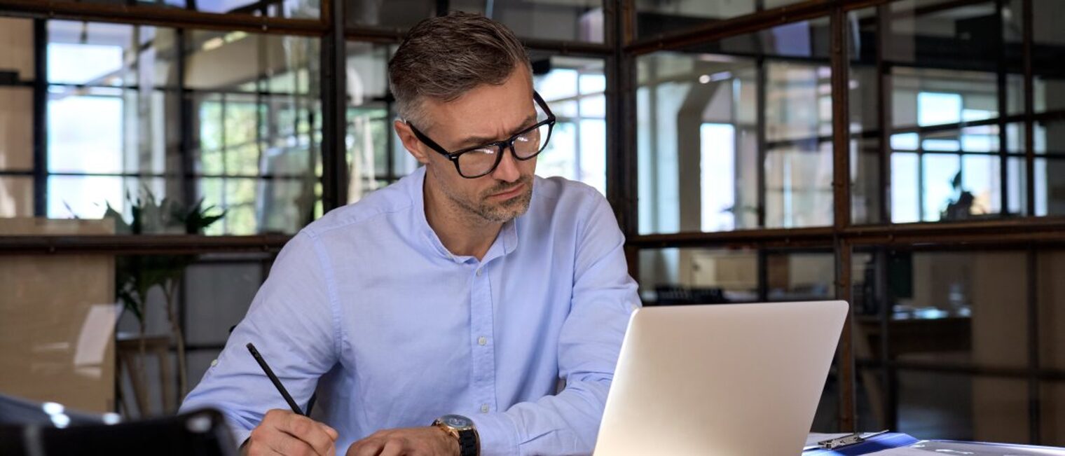 Ein Geschäftsmann sitzt im Büro am Laptop und nimmt an einem Web-Seminar teil, er macht sich Notizen.