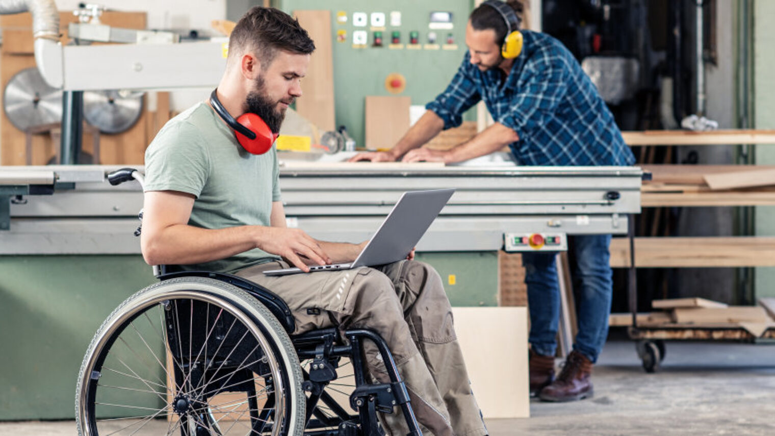In einer Schreiner-Werkstatt sitzt ein junger Mann im Rollstuhl, er hat einen Laptop auf dem Schoß liegen.