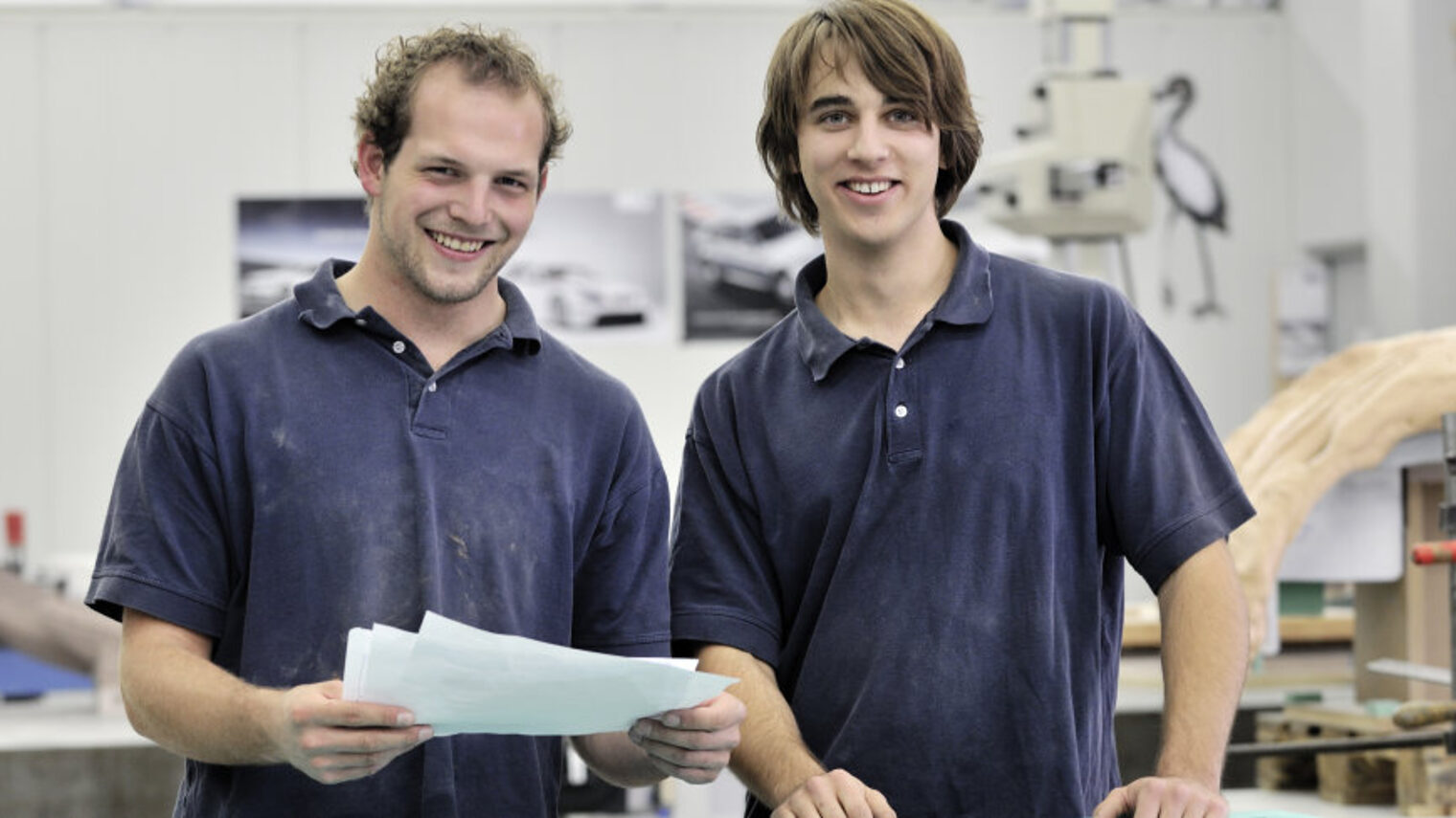 Zwei junge Handwerker stehen fröhlich nebeneinander in einer Werkstatt.