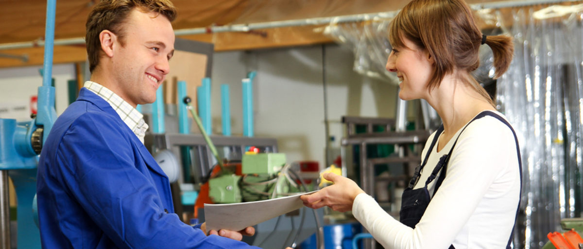 Mann in blauem Arbeitskittel reicht junger Frau in Latzhose lächelnd die Hand und einige Unterlagen in einer Werkstatt.