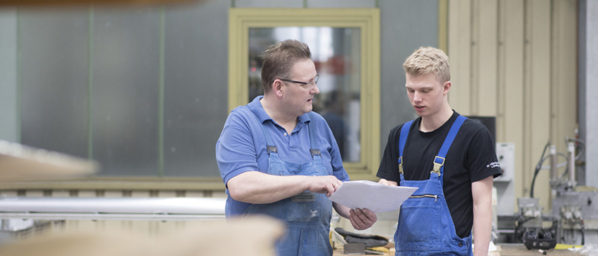 Ausbildungsordnung und Berichtsheft sind die Grundlagen einer Lehre. Zwei Männer in Arbeitskleidung stehen in einer Produktionshalle. Der Ältere zeigt dem Jüngeren ein Blatt Papier und spricht.