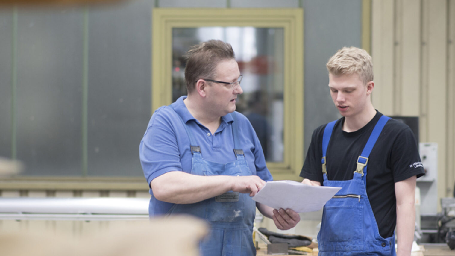 Zwei Männer in Arbeitskleidung stehen in einer Produktionshalle. Der Ältere zeigt dem Jüngeren ein Blatt Papier und spricht.