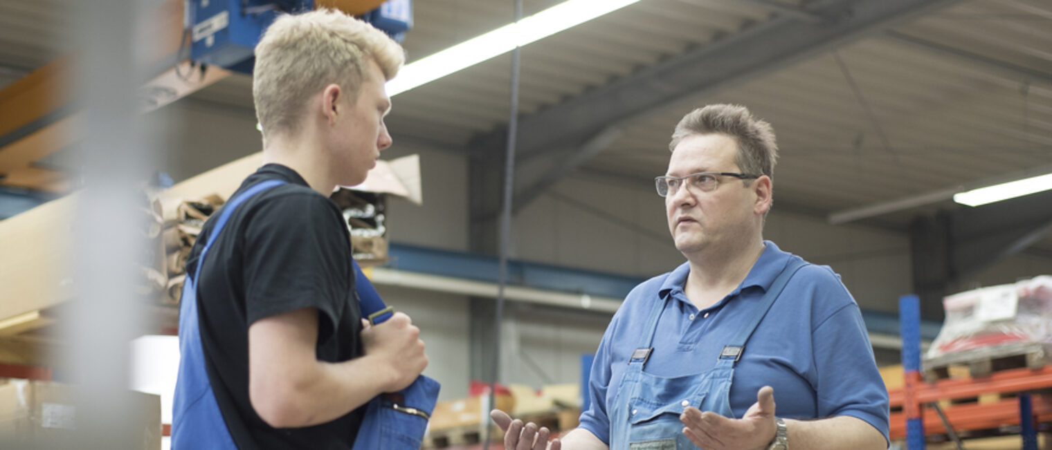 Konflikte in der Ausbildung mit einfachen Tipps vermeiden oder lösen. Ein Mann in Arbeitskleidung spricht mit einem jungen Mann in einer Lagerhalle. Dabei gestikuliert er mit den Händen.