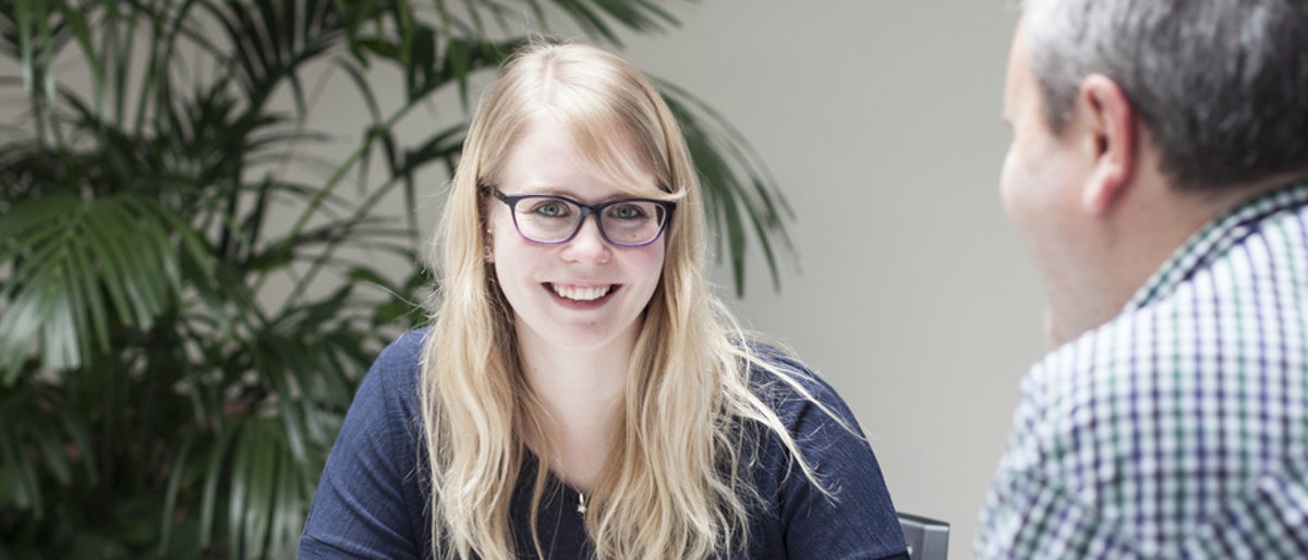 Einstellung neuer Mitarbeiter Ein Frau mit langen blonden Haaren und Brille lächelt in die Kamera. Rechts vorn im Bild sitzt ein Mann mit Rücken zur Kamera.