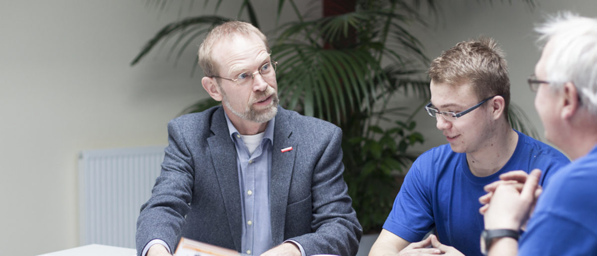 Förderprogramme unterstützen Betriebe bei der Ausbildung. Drei Männer sitzen an einem Tisch. Der Man im Sakko spricht zu den anderen beiden in blauen T-Shirts.