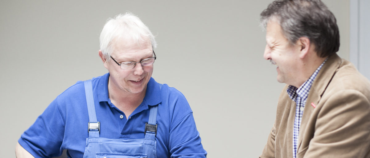 Handwerker können sich mit Fragen zur Ausbildung an die Berater wenden Ein Mann in blauem T-Shirt und Latzhose von vorne zu sehen, lacht. Ein Mann in Hemd und Jacket blickt ihn von der Seite an.