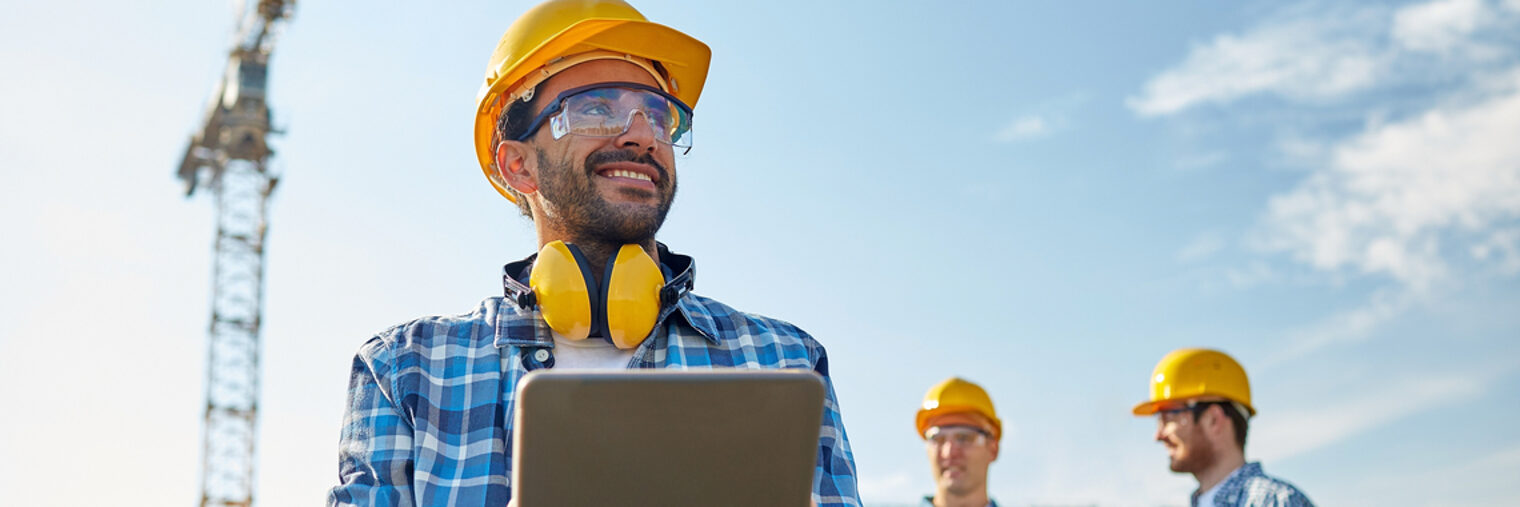 Drei Männer stehen mit Schutzhelm auf der Baustelle, im Hintergrund ein Kran. Der Mann im Vordergrund hält ein Tablet in der Hand.