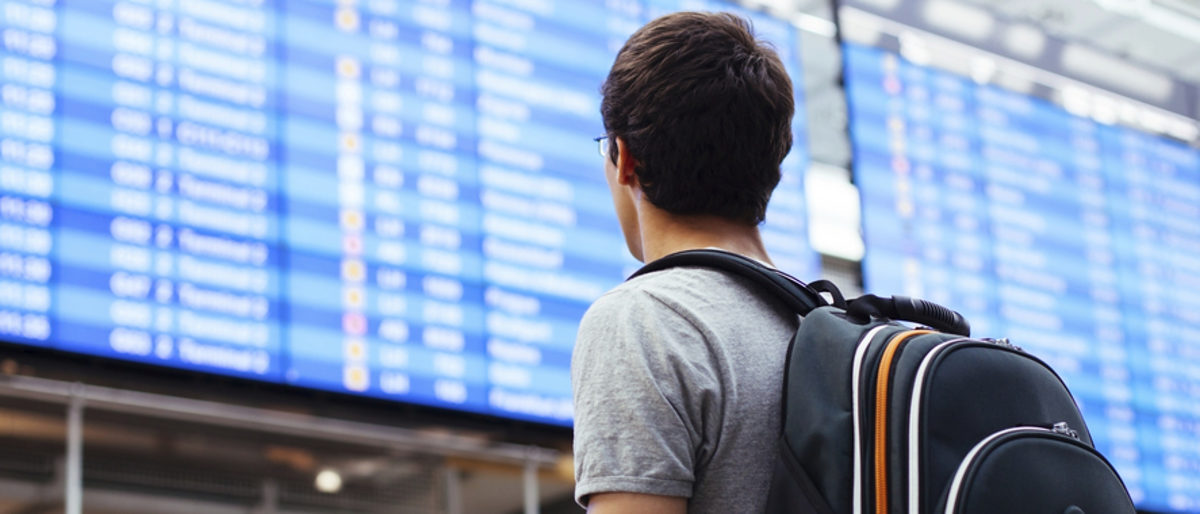 Azubis gewinnen durch Praktika im Ausland wertvolle Erfahrungen. Eine Person mit einem Rucksack ist von hinten zu sehen, im Hintergrund eine Anzeigentafel wie an Bahnhöfen oder Flughäfen.