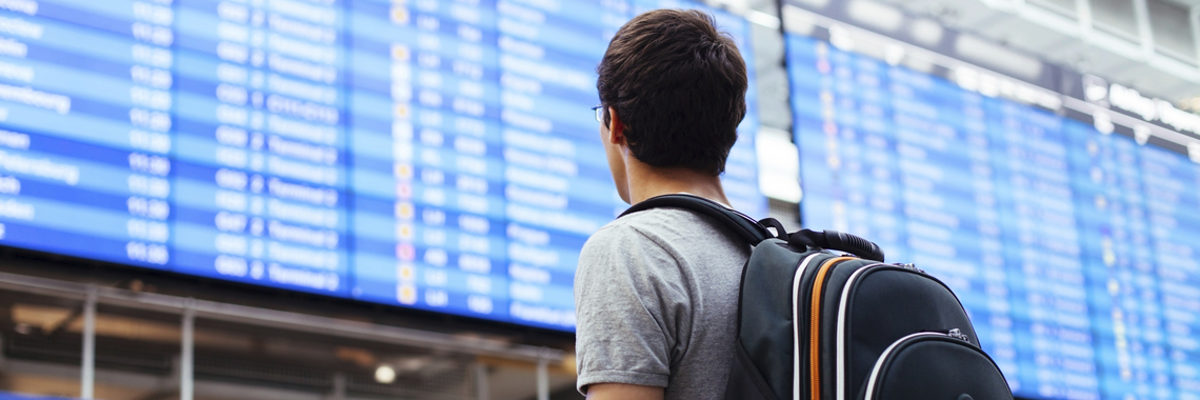 Azubis gewinnen durch Praktika im Ausland wertvolle Erfahrungen. Eine Person mit einem Rucksack ist von hinten zu sehen, im Hintergrund eine Anzeigentafel wie an Bahnhöfen oder Flughäfen.