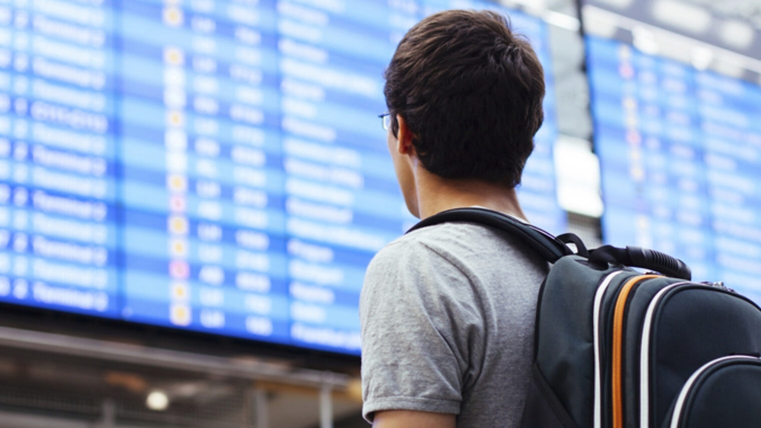 Eine Person mit einem Rucksack ist von hinten zu sehen, im Hintergrund eine Anzeigentafel wie an Bahnhöfen oder Flughäfen.