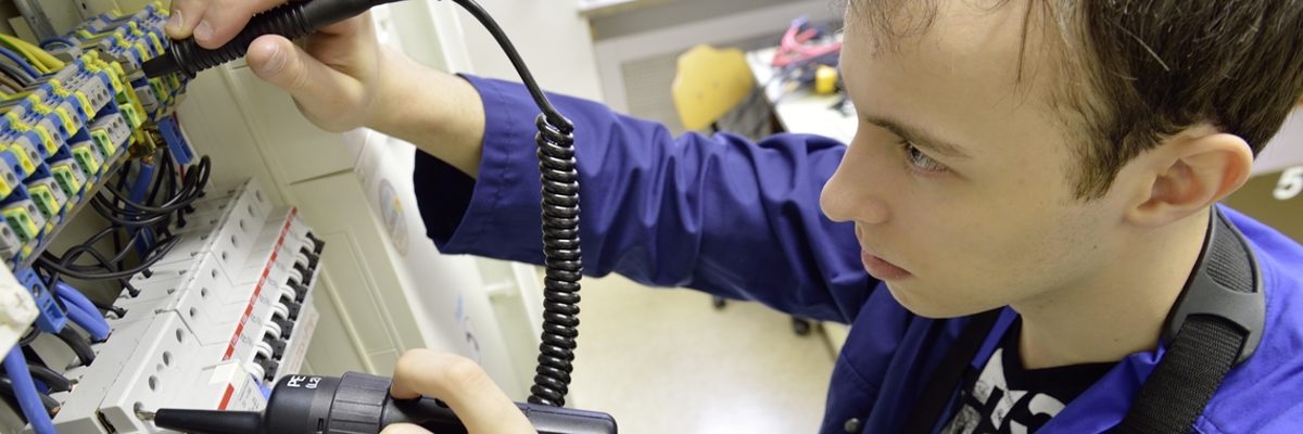 Beim Leistungswettbewerb zeigen junge Handwerker, was sie können. Ein junger Mann sieht konzentriert in einen Stromkasten. In beiden Händen hat er Werkzeuge zum Messen von Strom.