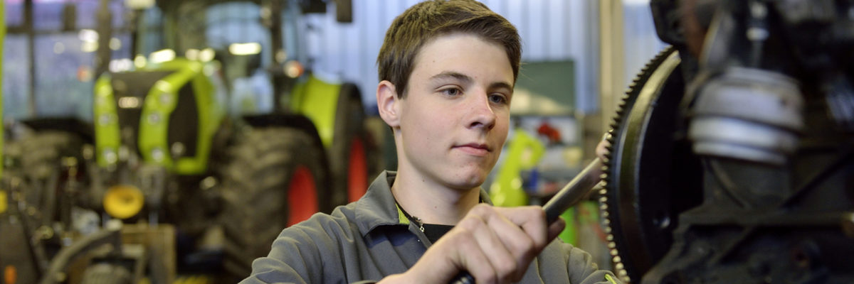 Die Zahlen zur Ausbildung im Handwerk der Region Heilbronn-Franken. Junger Mann zieht in einer Werkstatt mit Werkzeug eine Schraube an. Im Hintergrund sieht man unscharf einen Traktor.