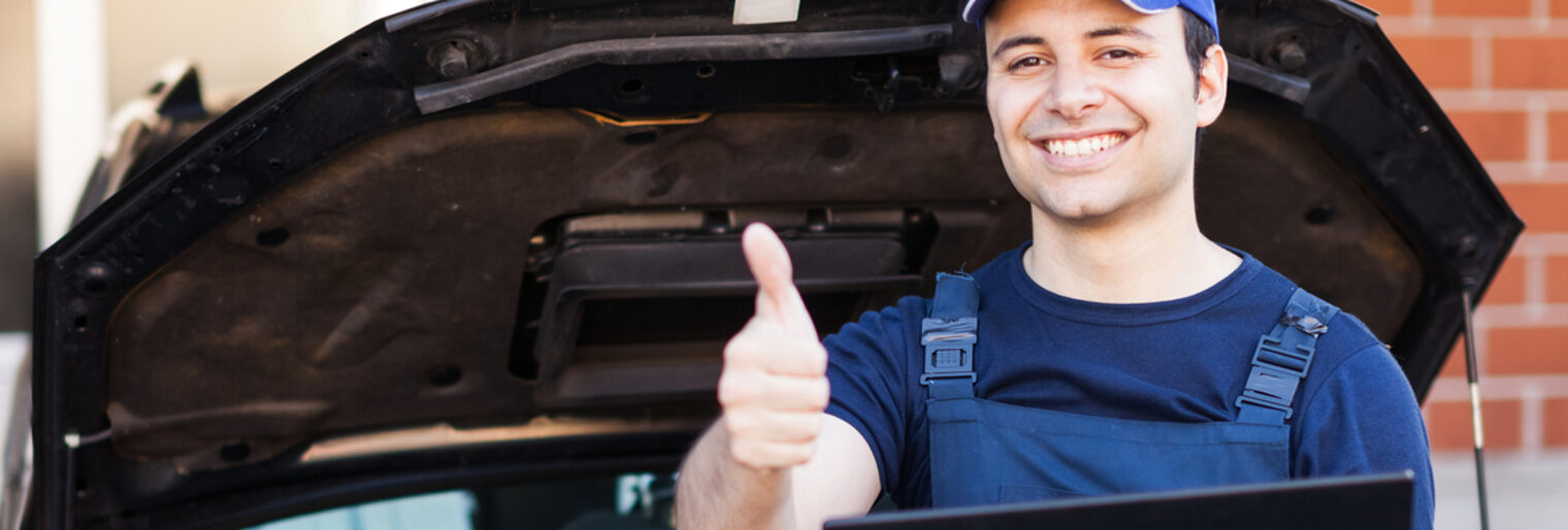 Kfz-Mechaniker lehnt an geöffneter Motorhaube mit Laptop, strahlend zeigt er mit der Hand den Daumen hoch.