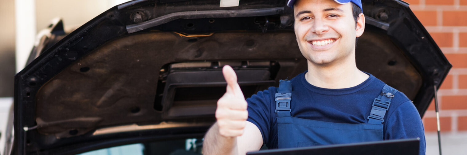 Kfz-Mechaniker lehnt an geöffneter Motorhaube mit Laptop, strahlend zeigt er mit der Hand den Daumen hoch.