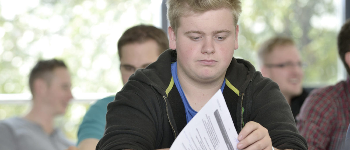 Informationen rund um die Meisterprüfung für angehende Meisterschüler. Junger Mann blättert in Unterlagen, im Hintergrund sind weitere Männer in einem Schulungsraum unscharf zu sehen.