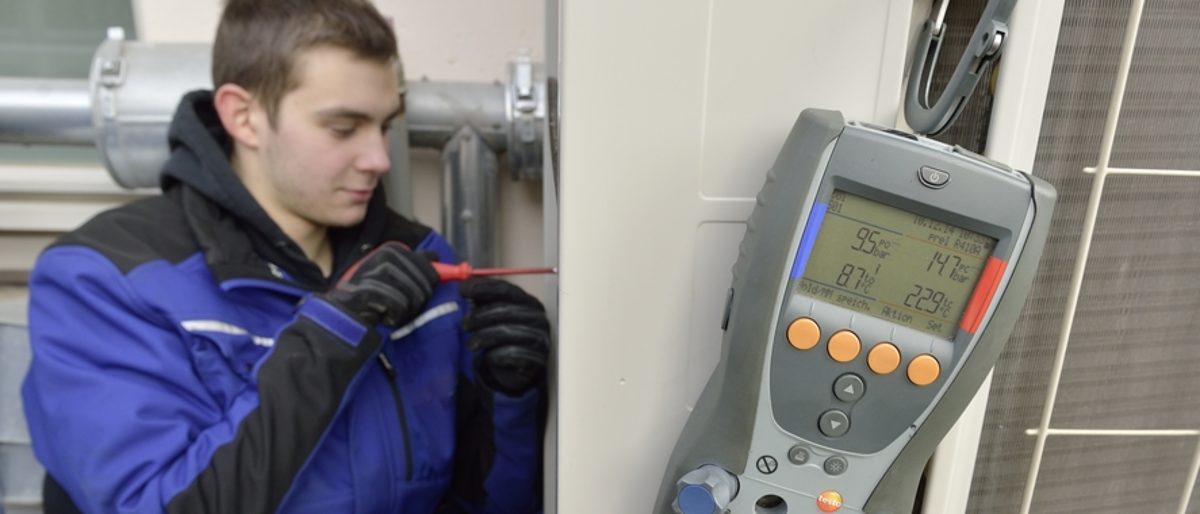 Mechatroniker für Kältetechnik ist einer von 130 Berufen im Handwerk. Im Vordergrund eine Messgerät. Im Hintergrund ein junger Mann in blau-schwarzer Arbeitskleidung mit einem Schraubendreher.
