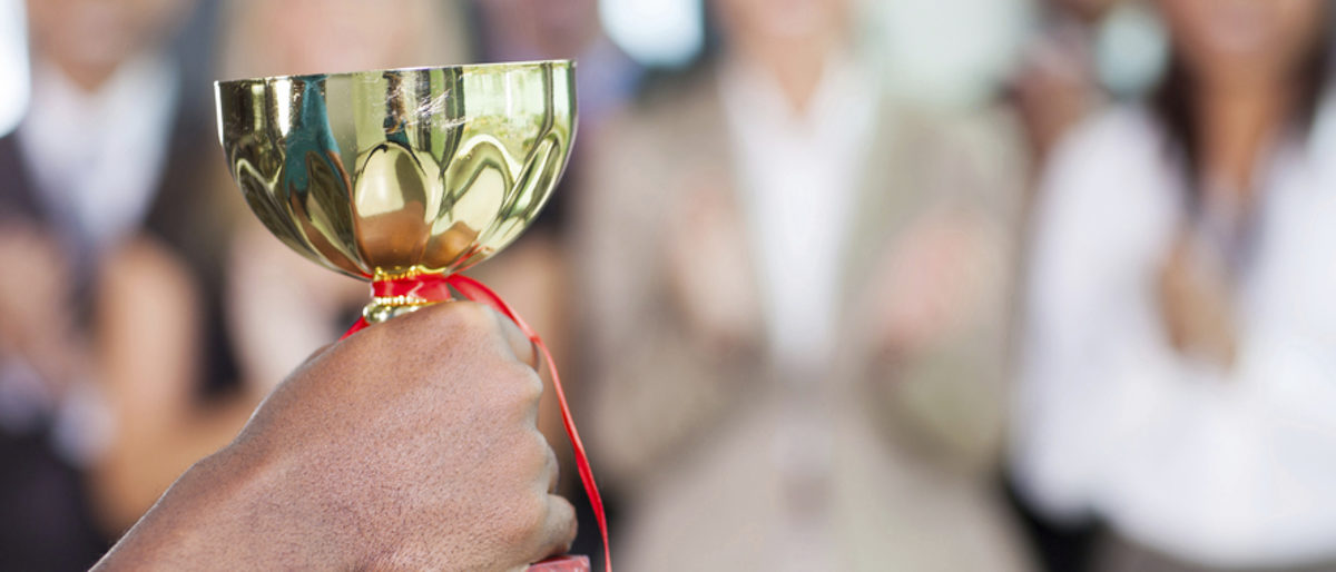 Wettbewerbe und Preise belohnen Betriebe für ihre Innovationen. Eine Hand hält einen goldenen Pokal in die Höhe, im Hintergrund applaudieren Menschen.