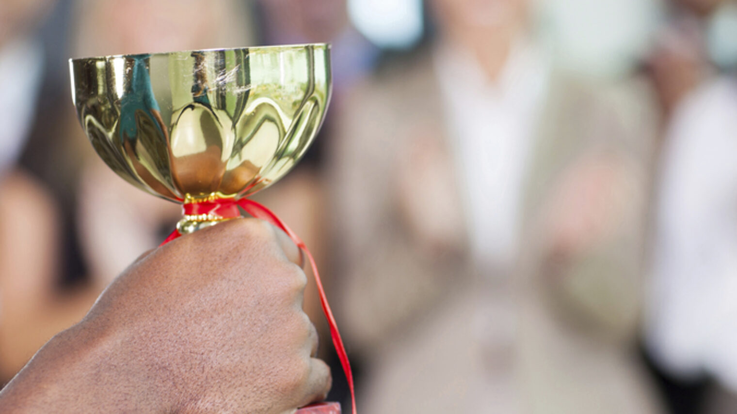 Eine Hand hält einen goldenen Pokal in die Höhe, im Hintergrund applaudieren Menschen.