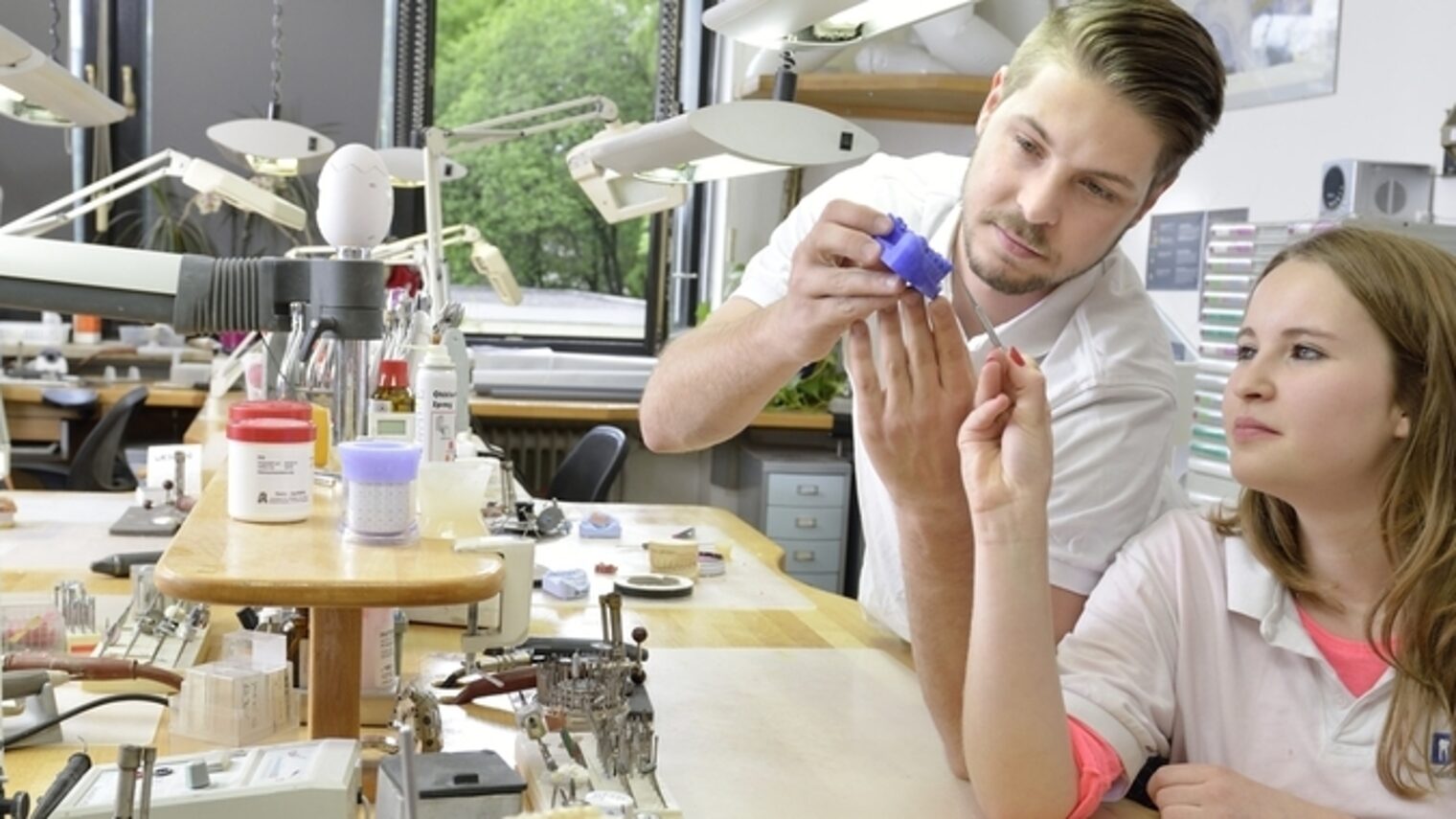 In einer Werkstatt sehen sich ein junger Mann und eine junge Frau einen blauen Zahnabdruck an. Die Frau hält ein Werkzeug.