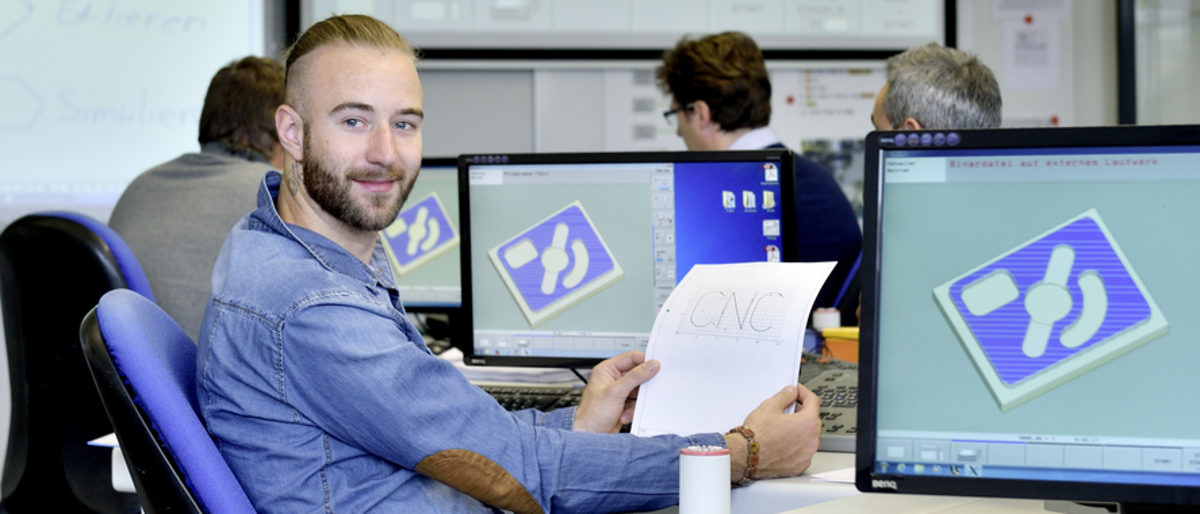 Statistik der Weiterbildungen für Handwerker in Heilbronn-Franken. Ein Mann sitzt zwischen zwei Computerbildschirmen. Er hält ein Blatt in der Hand und lächelt in die Kamera.