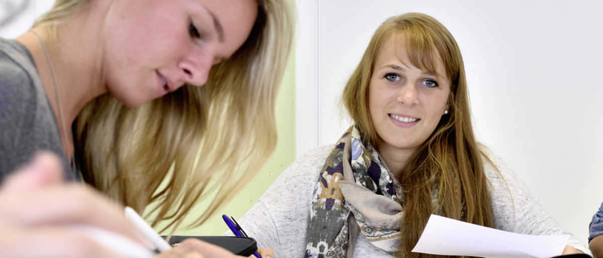 Ein Duales Studium bietet auch im Handwerk schnelle Aufstiegschancen. Eine junge Frau lächelt in die Kamera. Um Sie herum sieht man im Anschnitt weitere junge Menschen, die schreiben oder lernen.