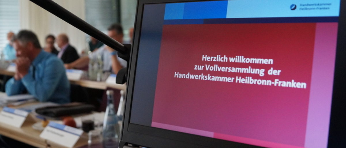 Viele Handwerker engagieren sich ehrenamtlich in verschiedenen Gremien Display mit "Herzlich willkommen zur Vollversammlung der Handwerkskammer Heilbronn-Franken", im Hintergrund sitzen Menschen.