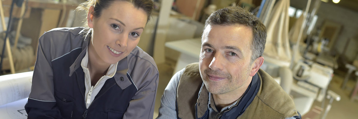 Das Allgemeine Gleichbehandlungsgesetz soll vor Diskriminierung schützen. Eine Frau und ein Mann in braun-grauer Arbeitskleidung lächeln in die Kamera. Im Hintergund ist eine Schreiner-Werkstatt zu sehen.