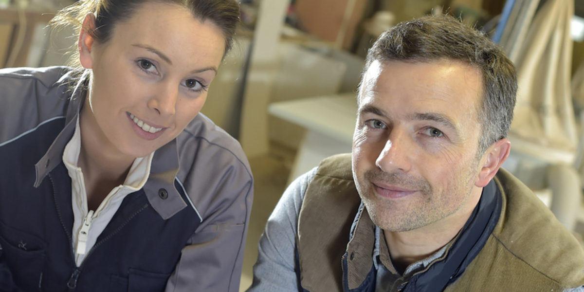 Eine Frau und ein Mann in braun-grauer Arbeitskleidung lächeln in die Kamera. Im Hintergund ist eine Schreiner-Werkstatt zu sehen.