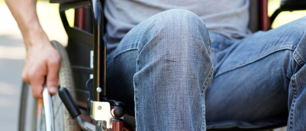 Für Schwerbehinderte gibt es besonderen Schutz, auch in der Arbeitswelt. Jemand, der eine Jeans trägt, sitzt im Rollstuhl. Der Oberkörper ist nicht zu sehen. Eine Hand greift ans Rad.