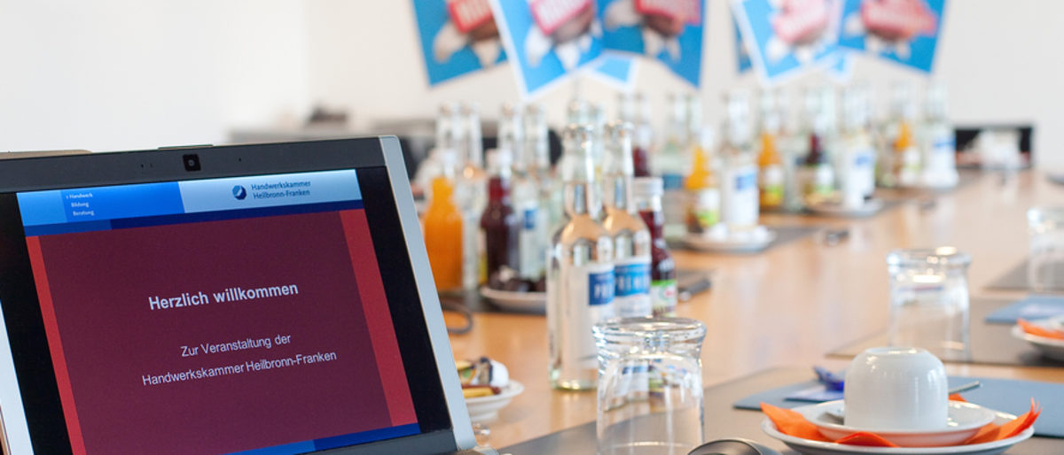 Der Berufsbildungsausschuss ist in wichtigen Bildungsfragen zu hören. Im Vordergrund der Bildschirm eines Laptops. Im Hintergrund ein großer Besprechungstisch mit Tassen, Gläsern und Getränken.