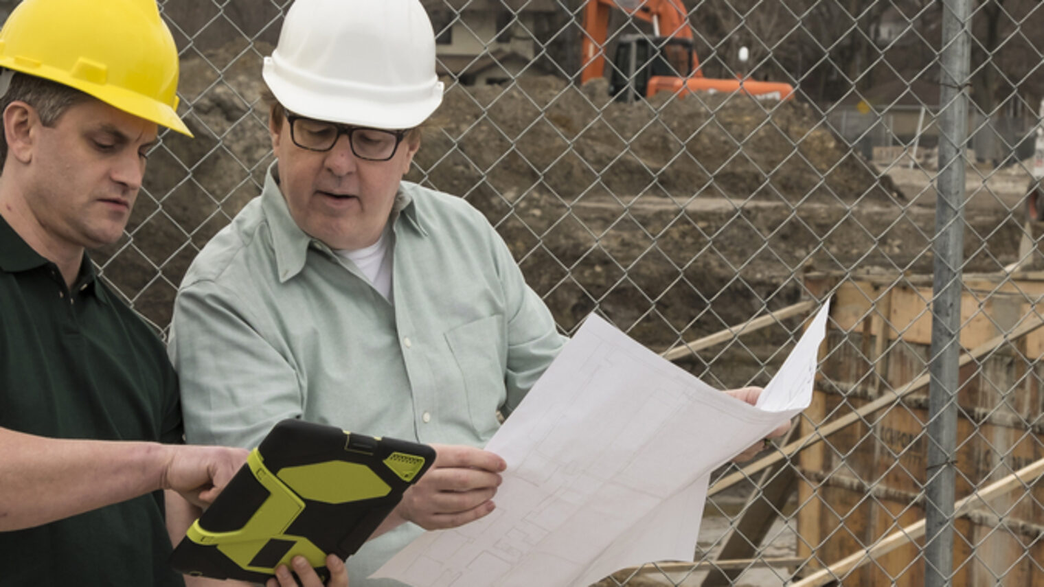 Zwei Männer mit Helmen schauen auf einen Plan. Im Hintergrund ist eine Baustelle zu sehen.